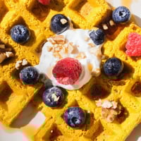 A stack of golden, fluffy Anti-Inflammatory Adaptogenic Turmeric Waffles topped with fresh blueberries and a drizzle of maple syrup.